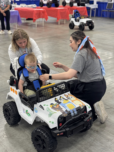 A young child sits in a toy car decorated with Bluey characters while two adults strap the child in.