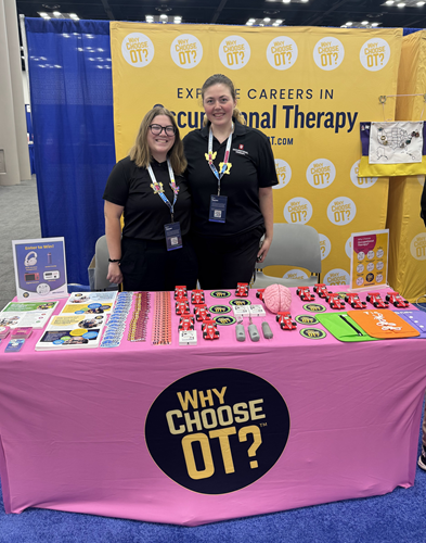 Two young women stand behind a table full of swag. The tablecloth is printed with Why Choose OT?