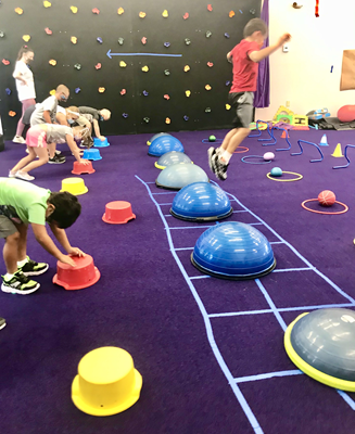 In a therapy gym, a child jumps over a Bosu ball while other children practice stretching.