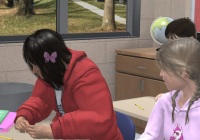 A young girl sits in a classroom, shoulders hunched, while her classmate looks at her.