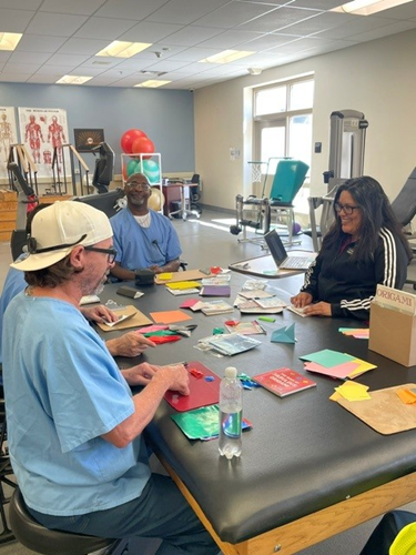 A woman and three men sit around a table folding paper. Colorful paper is strewn across the table.