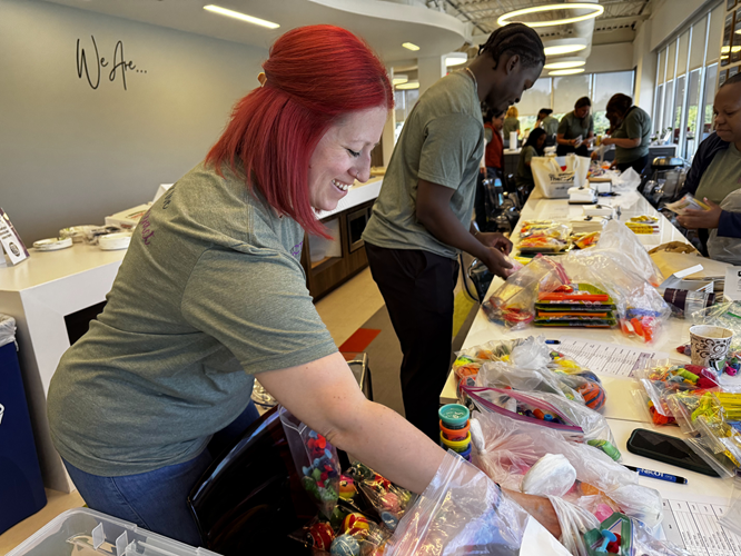 A red-haired woman in the foreground and other people in the background around a long table strewn with supplies.