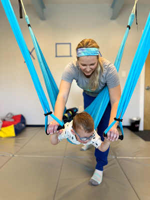 A child lies on his stomach on a sensory swing while a therapist supports him from above.