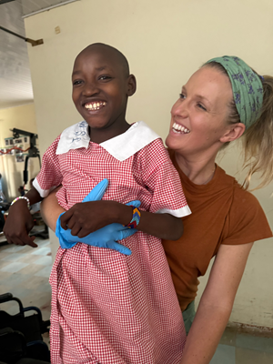 A young Black girl smiles wide for the camera while her therapist has an arm wrapped around the girl's waist for support.