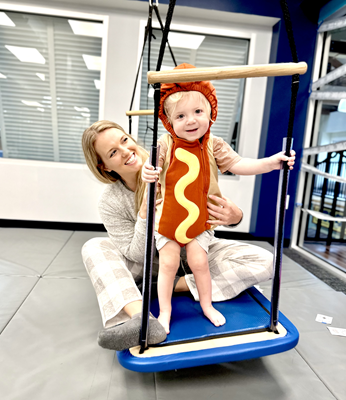 A child wearing a hot dog costume stands on a balance platform. A therapist sits behind the child, supporting them.