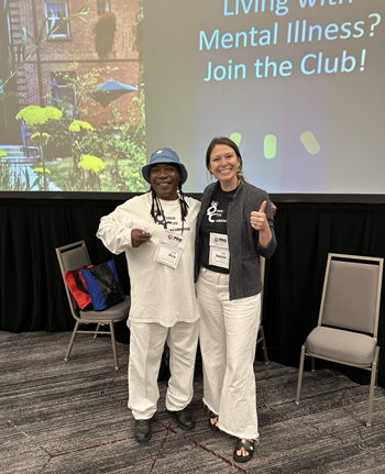 Two women pose in front of a projector screen that says "Living With Mental Illness? Join the Club!"