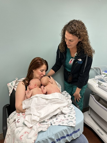 A woman reclined in a chair cradles two babies against her chest. An occupational therapist stands next to the woman offering her support.