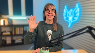 A woman sits in front a microphone and waves to the camera.